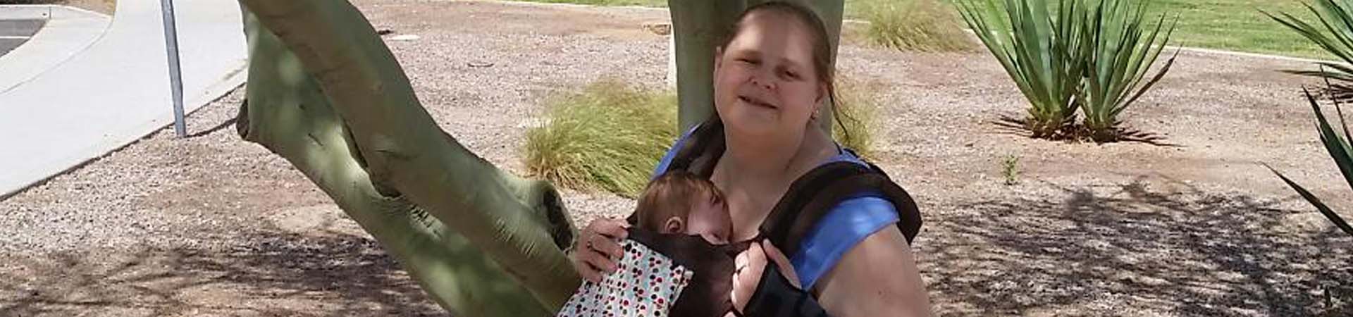 Blind mother smiles next to a tree, with her daughter, in a carrier, on her chest.